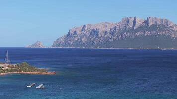 Sardinia's coastline features rugged cliffs, small islands, and a sailboat anchored in clear blue waters, with a mountain range in the background. video