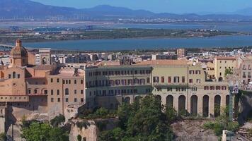 Cagliari's vibrant buildings on a rocky hill overlook blue waters and mountains, with wind turbines in the background, showcasing Sardinia's charm. video