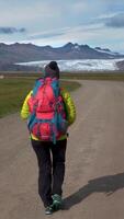 Female tourist with backpack goes by dirt road on background of mountains and glacier in Iceland. Freedom and travel concept. vertical footage video