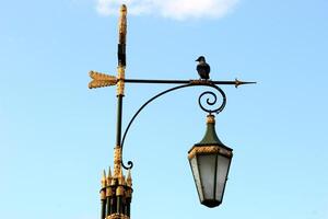 A close-up of an ornate lantern post featuring golden details and a bird perched on top. The lantern's intricate design contrasts beautifully against a clear blue sky photo