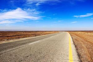 A scenic highway cutting through the arid desert landscape of Jordan, stretching endlessly into the horizon under a vast blue sky with scattered clouds. photo