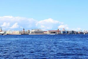Panoramic view of the Spit of Vasilyevsky Island with historic buildings and Rostral Columns in Saint Petersburg, Russia, against a blue sky and Neva River in the foreground. photo