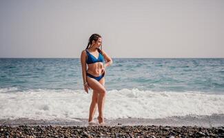 A tourist woman in blue bikini on beach enjoys the turquoise sea during her summer holiday. Rear view. Beach vacation. woman with braids dreadlocks standing with her arms raised enjoying beach ocean. photo
