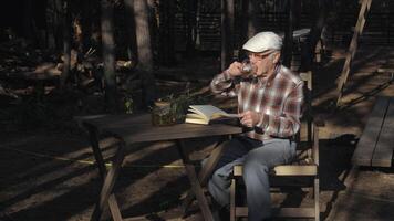 An elderly man sits at a wooden table in a forest setting, sipping tea from a mug while reading a book under the soft afternoon light, surrounded by nature. video
