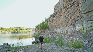 adult tourist is walking along the bank of the lake with a stick in his hand and with a backpack on his shoulders on the background of high rocks in the summer video