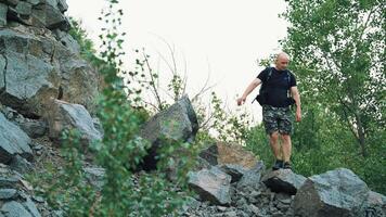 tourist in dark shorts and a t-shirt with a backpack on his shoulders is standing on the rocks and looking at the environment around himself on the background of green trees video
