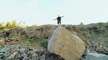 happy traveler with a backpack on his shoulders is standing at the top of a cliff with his arms raised and looking at the beautiful nature from high. Camera motion back video