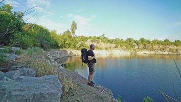 traveler in shorts and a black T-shirt with a backpack on his back is looking at the beautiful view from a cliff by the river in the summer video