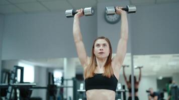 Pretty woman in black top raises up dumbbells on the background of the gym. Exercises for biceps. Close-up video