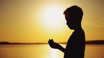 A little boy is making a paper boat with his own hands and playing with it in the air at sunset near the river in warm weather. video