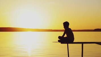 a little boy is sitting in a lotus position on the edge of a bridge above a river at sunset in the summer. video