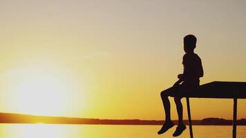 A little child is sitting on the bridge above the lake and throwing stones into the water at sunset in the summer on a warm evening. video