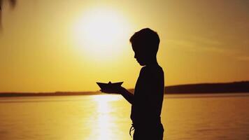 A little boy is holding an origami in the form of a paper boat in his hands and raising up it to the level of the sun on the background of the lake. Silhouette of a guy at sunset video