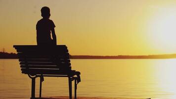 A little boy is looking at the sunset and wonderful landscape near the river on the bench. A guy raises his hands up. Weekend. video
