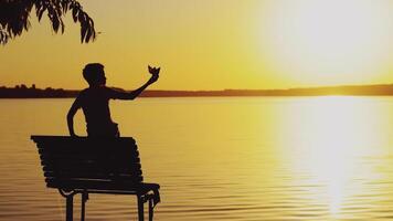 a little boy is sitting on the bench and playing with a paper boat on the background of evening landscape by the lake in the summer. Sunset video
