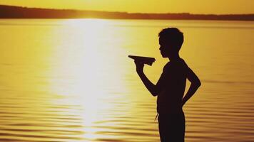 A little boy is holding a paper airplane in his hand and playing with him on the bank of the river at sunset on the background of the solar track. video