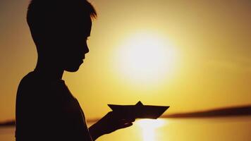 Silhouette of a little boy is holding a paper boat in his hand and looking at it on the background of sunset in the summer near the pond. Close-up video