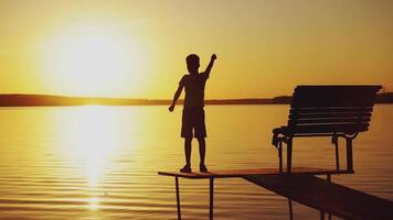 A little boy is standing on the masonry near the bench and doing physical exercises. The child is raising hands up alternately on the background of the river at sunset. video