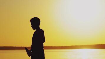 little kid holds origami of paper airplane in his hand and plays at sunset on the background of lake. A boy passionates about playing with his paper toy video