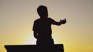 boy is holding paper ship in his hand and playing with it on the background of sunset by the river in the summer video