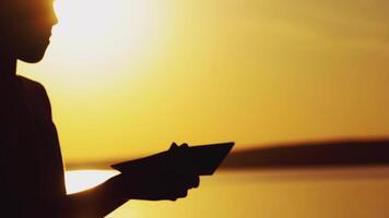 boy is playing with origami of handmade in the form of a boat on the background of sunset by the river in the summer video