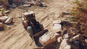 professional equipment with a bucket is turning over a stone and raising it near sandy quarry on the background of trees. Close-up video
