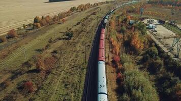 a long freight train with building materials is moving by railway on the background of fields and forests in the summer on a sunny day. Aerial view video