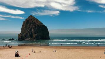 Giant Haystack Rock formation on a sandy beach with visitors. A towering rock formation stands tall on a sandy beach as visitors walk along the shore. video