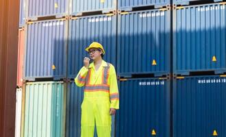 Asian man worker talking on walkie talkie for control loading containers box while standing in front of blue containers at cargo shipping yard photo