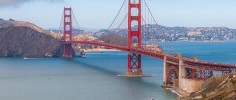 The Golden Gate Bridge spans the San Francisco Bay, its orange color contrasting with blue waters and distant hills. Sailboats add scale and activity. photo