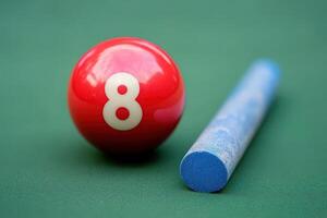 A vibrant red snooker ball marked with the number 8 rests next to a blue chalk stick on a smooth green felt surface. photo
