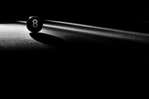 A lone snooker ball sits precariously at the edge of a dimly lit table, casting a shadow. The ambient light creates a dramatic effect, enhancing the balls colors and textures. photo