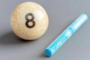 A solitary snooker ball rests next to a piece of blue cue chalk on a smooth gray surface, emphasizing the essentials of cue sports in a minimalist setting ideal for focusing on the game. photo