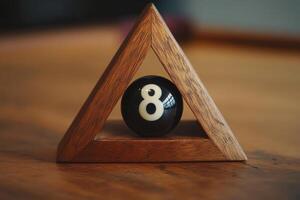 A black snooker ball with the number eight sits alone within a wooden triangle rack on a polished table, creating a quiet moment in a gaming environment. Soft light highlights the scene. photo