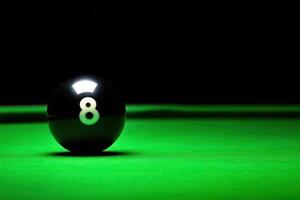 A single black snooker ball rests precariously on the edge of a green table, surrounded by a shadowy atmosphere. The low lighting highlights the glossy surface and the number eight. photo