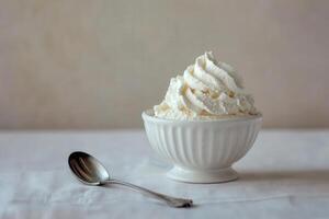 A small bowl holds fluffy whipped cream with a delicate texture, resting on a white tablecloth beside a shiny spoon. The minimalist setting emphasizes the delicious treat. photo