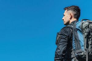 A border control officer is stationed outdoors, looking towards the horizon while wearing a backpack. The bright blue sky serves as a backdrop, highlighting the officers vigilant stance. photo