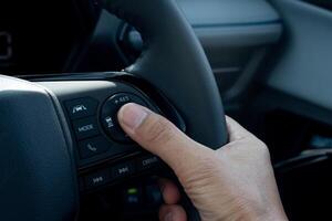 Hand of driver pressing a button on a car steering wheel. Steering wheel is black and has a leather or rubber grip. Hand is position in a way suggests the driver is using their thumb to press button. photo