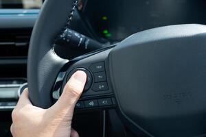 Hand of driver pressing a button on a car steering wheel. Steering wheel is black and has a leather or rubber grip. Hand is position in a way suggests the driver is using their thumb to press button. photo