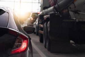 Traffic jam with several vehicles lined up on a road. Close up car with its taillights illuminated. Front of the car semi-trailer truck is partially visible. Sun is setting in the background. photo