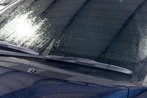 Close-up of a car windshield wiper. Wiper blade is black and appears to be made of rubber. Wiper arm is also black and has a metal spring attached to it. Background is blurred, of the hood of the car. photo