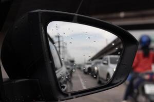 Side view mirror car reflecting a busy road with heavy traffic. Blurry and out of focus suggesting that the car is moving or that the mirror is dirty. On the mirror glass with drop of rain. photo