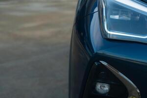 Front left corner of a blue luxury car. Car headlight is partially visible with its sleek and modern design shining through. Bumper is also visible with a slight curve. photo
