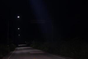 Empty road at night. Only light comes from a few street lamps that are illuminating the road ahead. Road is surrounded by trees that are casting long shadows. Scene appears to be quiet and peaceful. photo