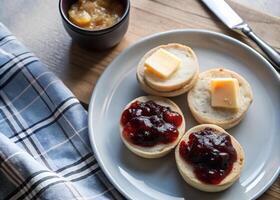 Top view of golden English muffins on a plate, one with butter melting, and the other spread with colorful jam. Soft morning light for a comforting breakfast scene photo