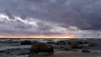 A breathtaking view of the sunset over the sea, with waves crashing against large boulders along the sandy shore. Captured from a low angle at eye level, highlighting the dramatic contrast video