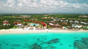 Aerial view of bright sunny day illuminates tropical beach of luxurious resort. People seen swimming and sunbathing on beach, which is dotted with palm trees and lounge chairs under thatched umbrellas video