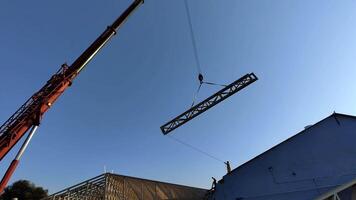 Construction workers guiding a large metal truss into place using a crane on a building site under a clear blue sky, with visible wooden framing of the structure video