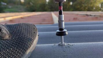 Close-up of a drill driving a screw into a corrugated roofing sheet during construction with a worker's shoe in the background video