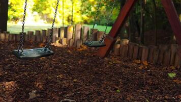 Empty black swing on children playground in the park in autumn season. Missing child. Lonely swing in sunny autumn day. Golden foliage in late fall. Beautiful nature. video